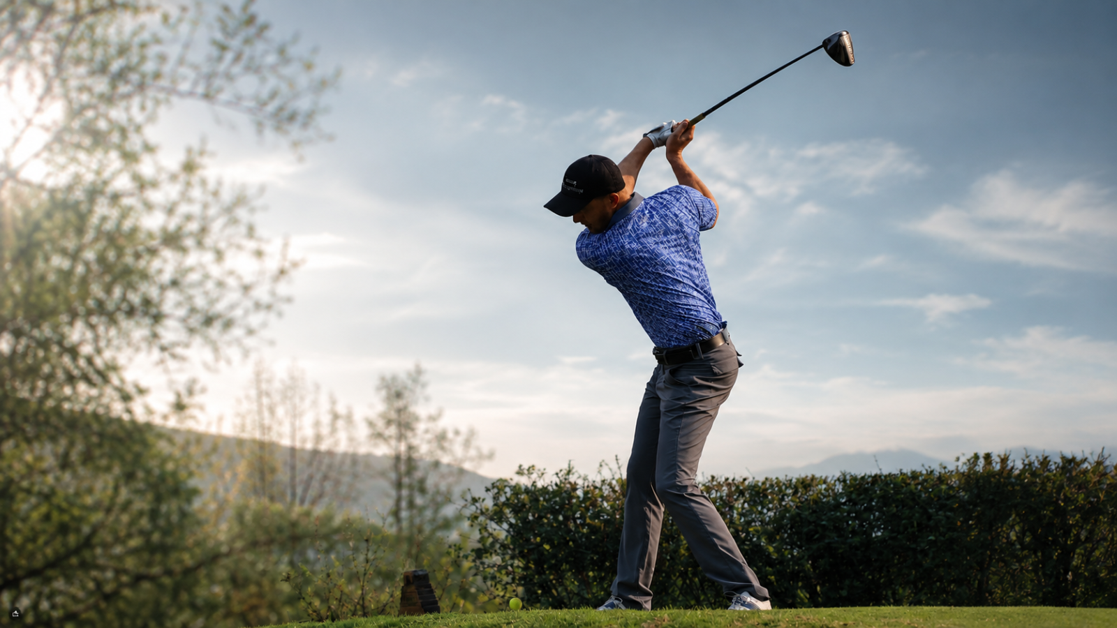 A male golfer mid-swing on a golf course, wearing a blue patterned DTWOOO performance polo shirt and dark pants.